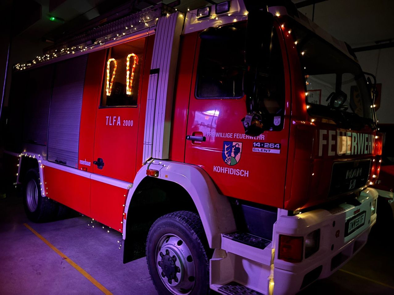 Ein roter Feuerwehrwagen mit beleuchteten Buchstaben und Zahlen steht in einer Garage, mit dekorativen Lichtern an der Seite und einem beleuchteten Fenster.