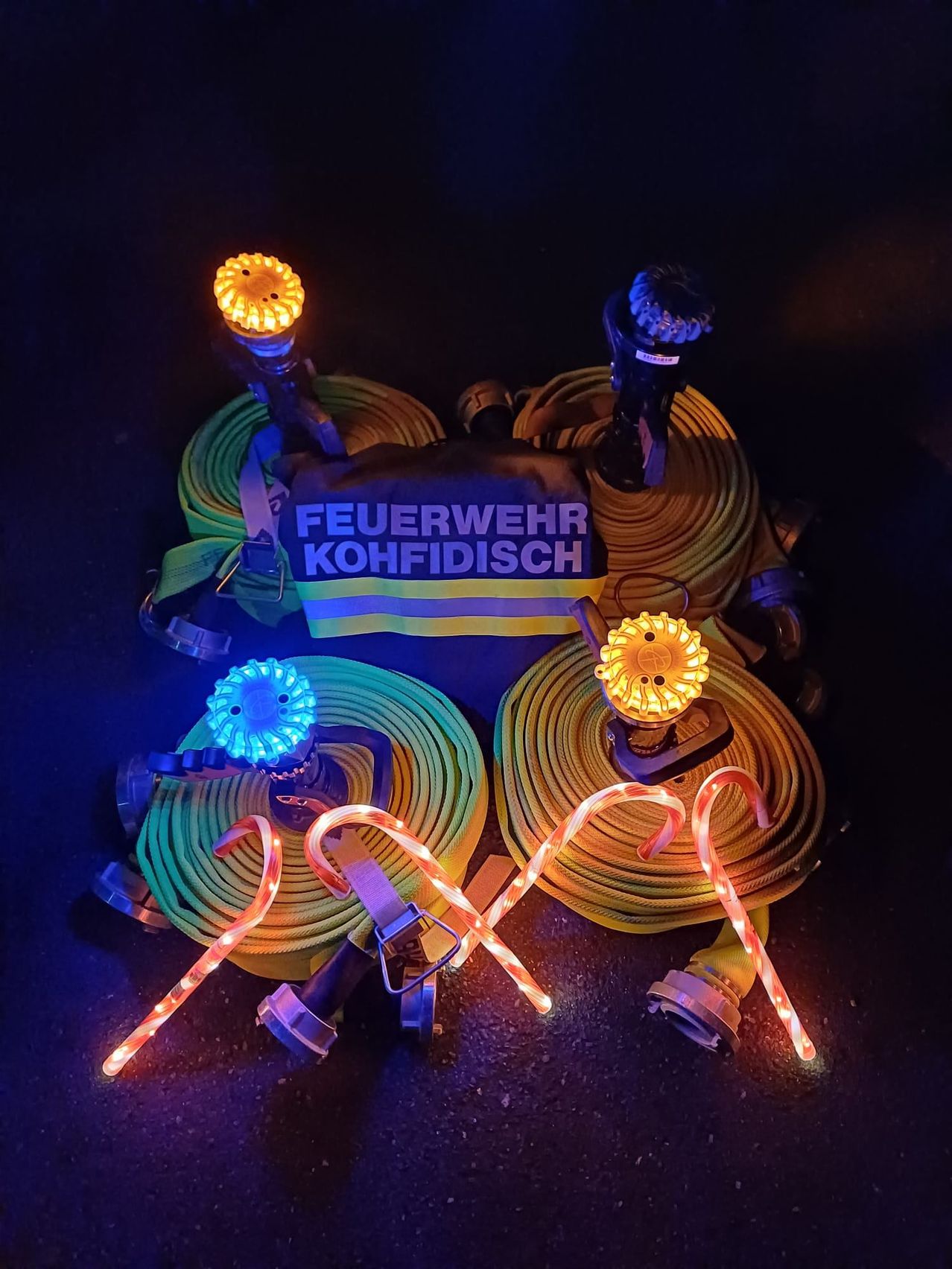 Eine Ausstellung von Feuerwehrausrüstung mit bunten Lichtern und den Worten 'Feuerwehr Kohfidisch' auf einem Stoff. Die Ausrüstung umfasst Feuerwehrschläuche mit dekorativen Weihnachtslichtern.