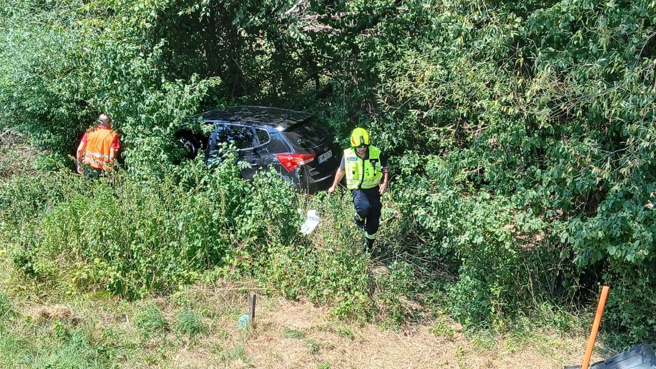Eine Person in einer gelben Weste und Helm läuft durch einen dichten Wald, möglicherweise auf der Suche nach etwas. Ein schwarzer SUV ist teilweise hinter ihr sichtbar.