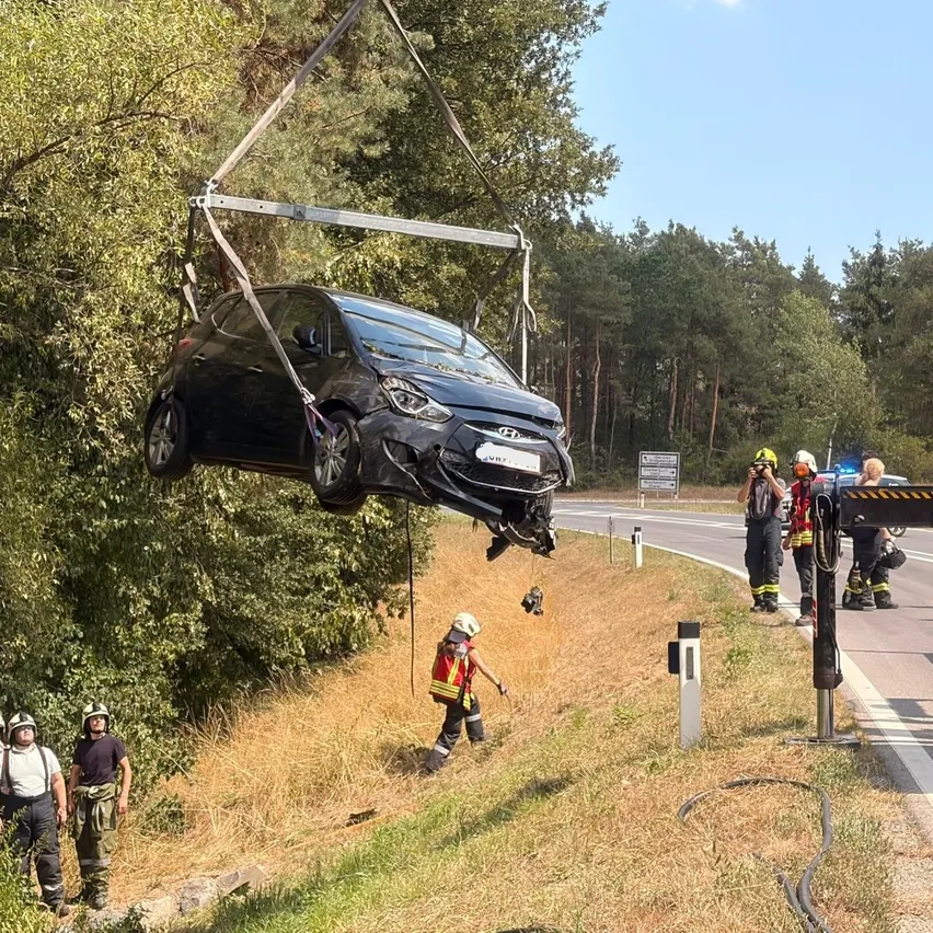Ein schwarzes Auto wird von einem Kran gehalten, mit Einsatzkräften am Straßenrand. Das Auto wird von einem Metallrahmen angehoben, und mehrere Arbeiter tragen Helme und Schutzausrüstung.