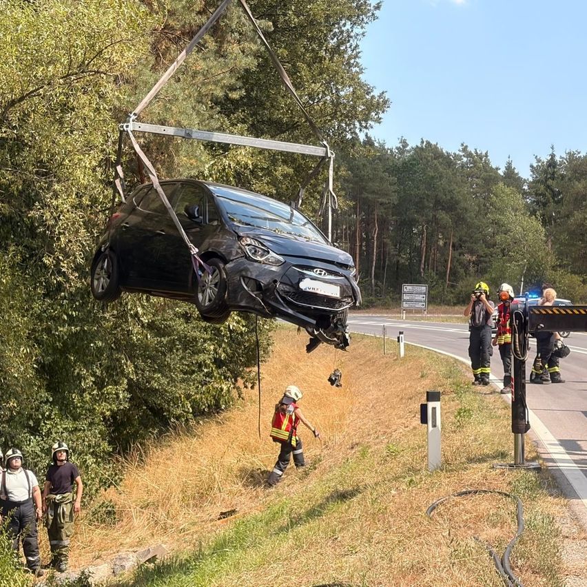 Ein schwarzes Auto wird von einem Kran gehalten, mit Einsatzkräften am Straßenrand. Das Auto wird von einem Metallrahmen angehoben, und mehrere Arbeiter tragen Helme und Schutzausrüstung.