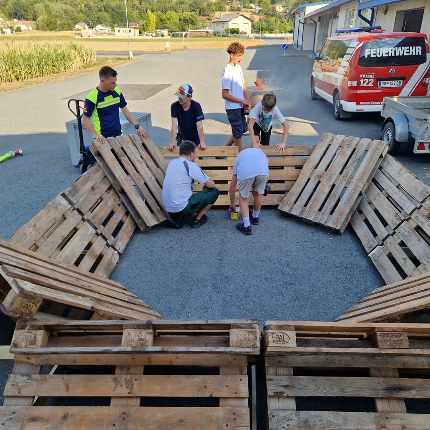 Eine Gruppe junger Menschen baut gemeinsam eine Struktur aus Holzpaletten auf einem gepflasterten Bereich. Ein Feuerwehrwagen ist in der Nähe geparkt.