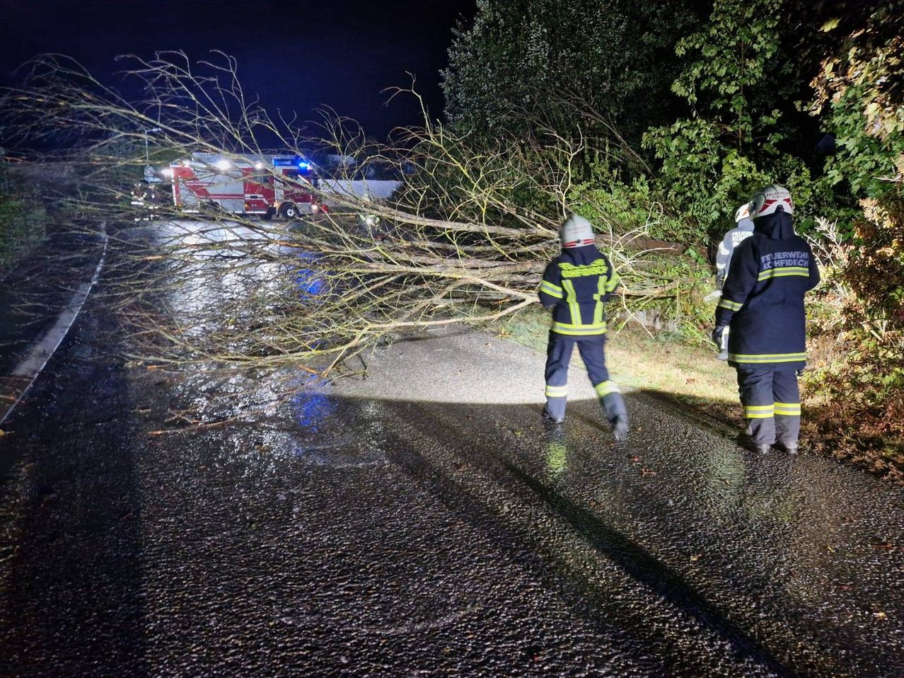 Zwei Feuerwehrleute gehen bei Nacht auf einer nassen Straße, mit einem umgestürzten Baum, der den Weg blockiert. Ein Feuerwehrauto ist in der Nähe geparkt.