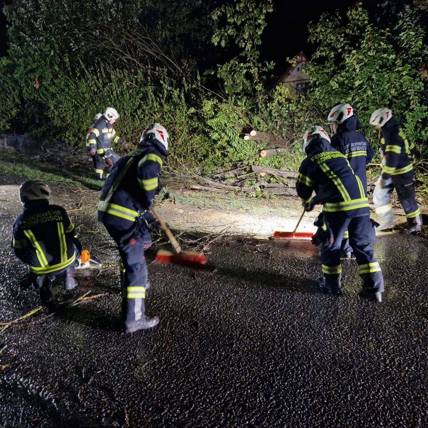 Eine Gruppe Feuerwehrleute räumt nachts Trümmer von einer Straße. Sie tragen Helme und Uniformen. Einige benutzen Besen und andere haben Kettensägen.