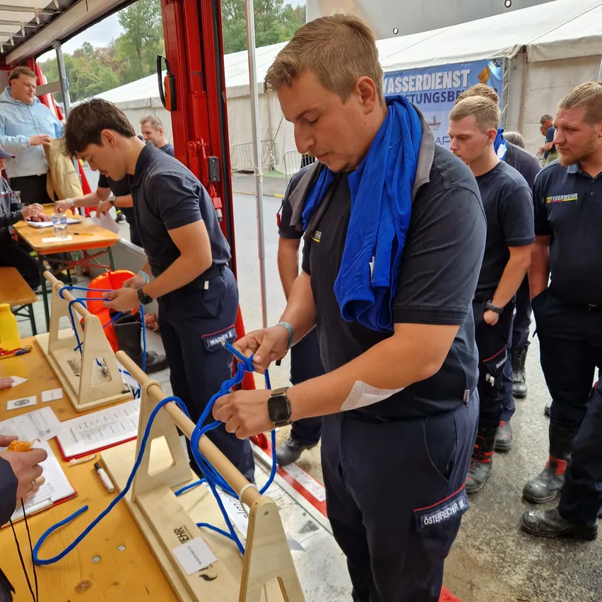 Eine Gruppe von Feuerwehrleuten übt das Knotenlegen mit Seilen an einem Holzrahmen. Sie tragen Uniformen und einige haben blaue Schals. Ein Tisch mit Papieren und einem Eimer ist in der Nähe.