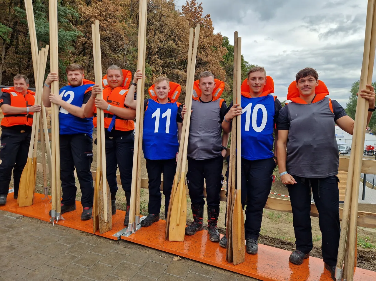 Fünf Männer stehen in einer Reihe, jeder hält eine Paddel und trägt Rettungswesten mit den Nummern 12, 11, 10 und zwei graue Westen. Sie stehen auf einer orangefarbenen Plattform mit einem Holzzaun im Hintergrund.