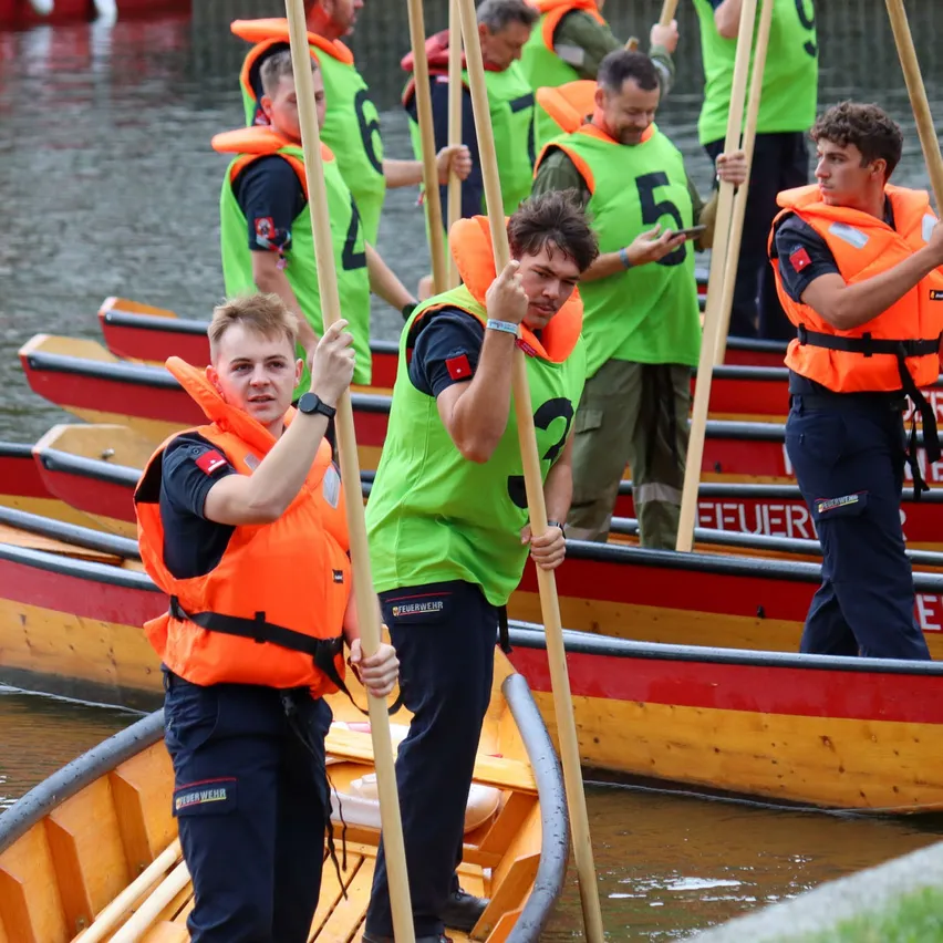 Eine Gruppe von Personen in grünen und orangefarbenen Rettungswesten steht in Booten, hält Paddel und nimmt möglicherweise an einer Wassersportveranstaltung teil.