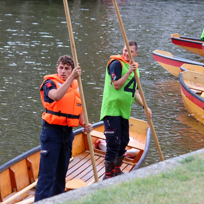 Zwei junge Männer in Rettungswesten paddeln ein Holzboot auf einem ruhigen Fluss, wobei andere Boote im Hintergrund sichtbar sind.