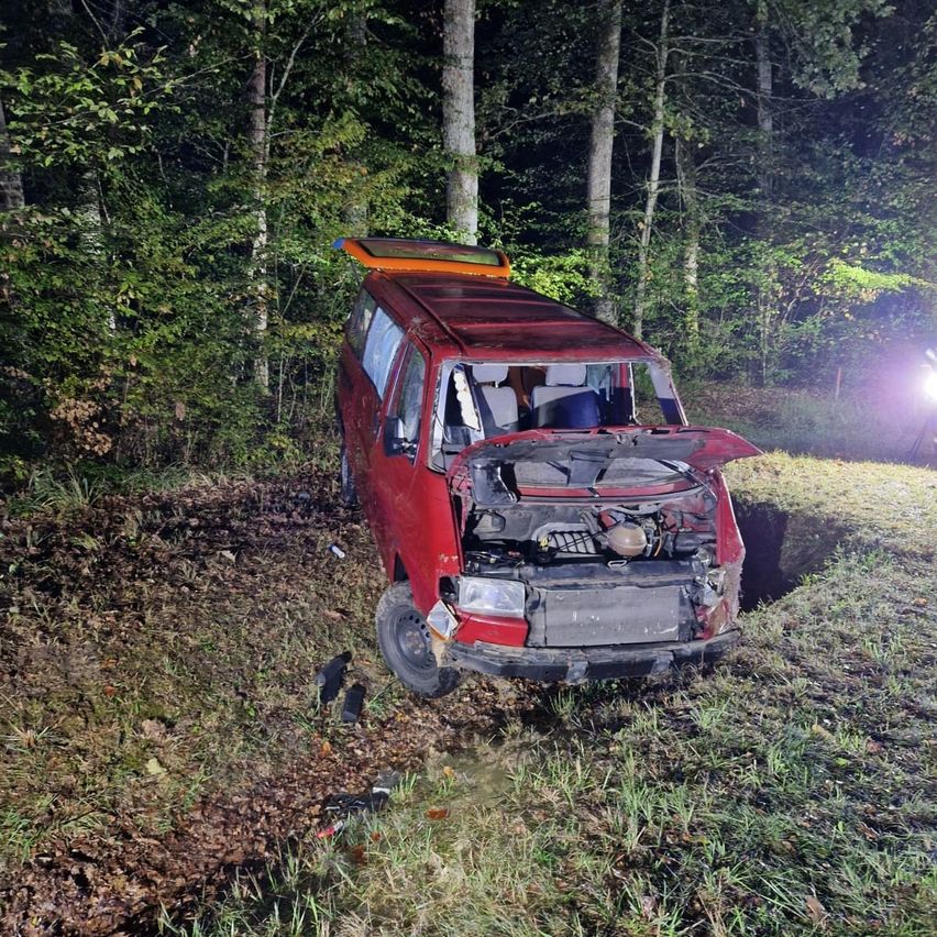 Ein roter Van ist in einem Wald bei Nacht verunglückt, mit stark beschädigter Front und offener Motorhaube.