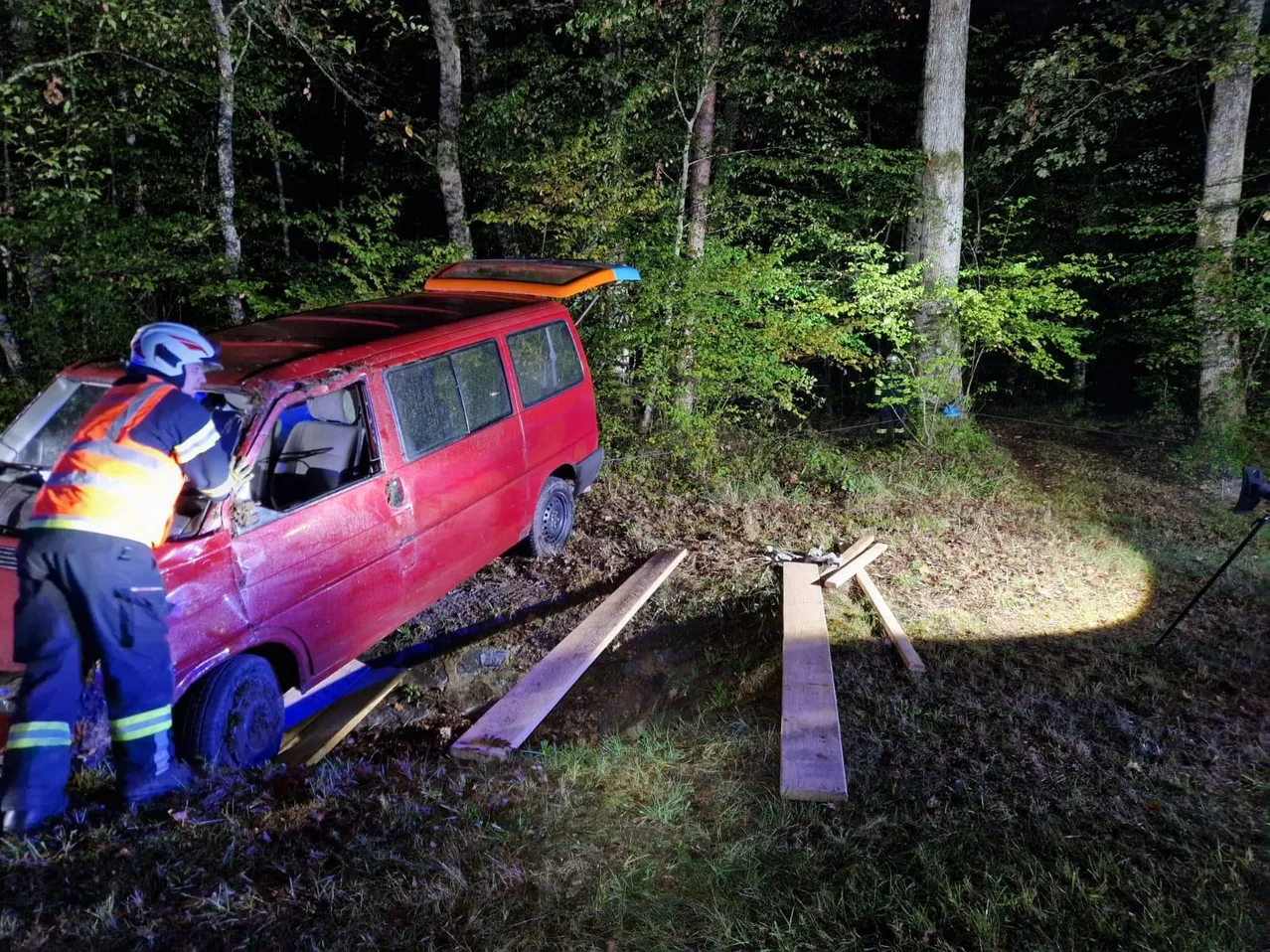 Ein roter Van ist in einem Wald verunglückt, mit offener Heckklappe und einem Feuerwehrmann in der Nähe.