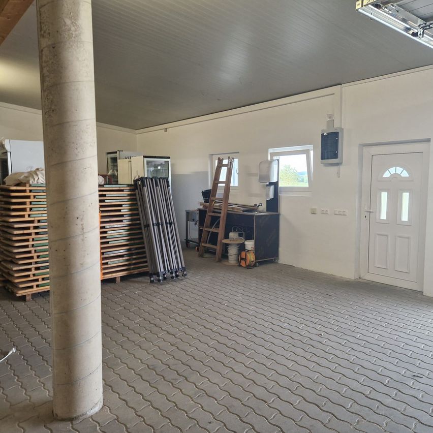 An empty room with tiled floor, a large pillar, wooden pallets, a ladder, and a refrigerator. The walls are white, and there is a window with a view of the outdoors.
