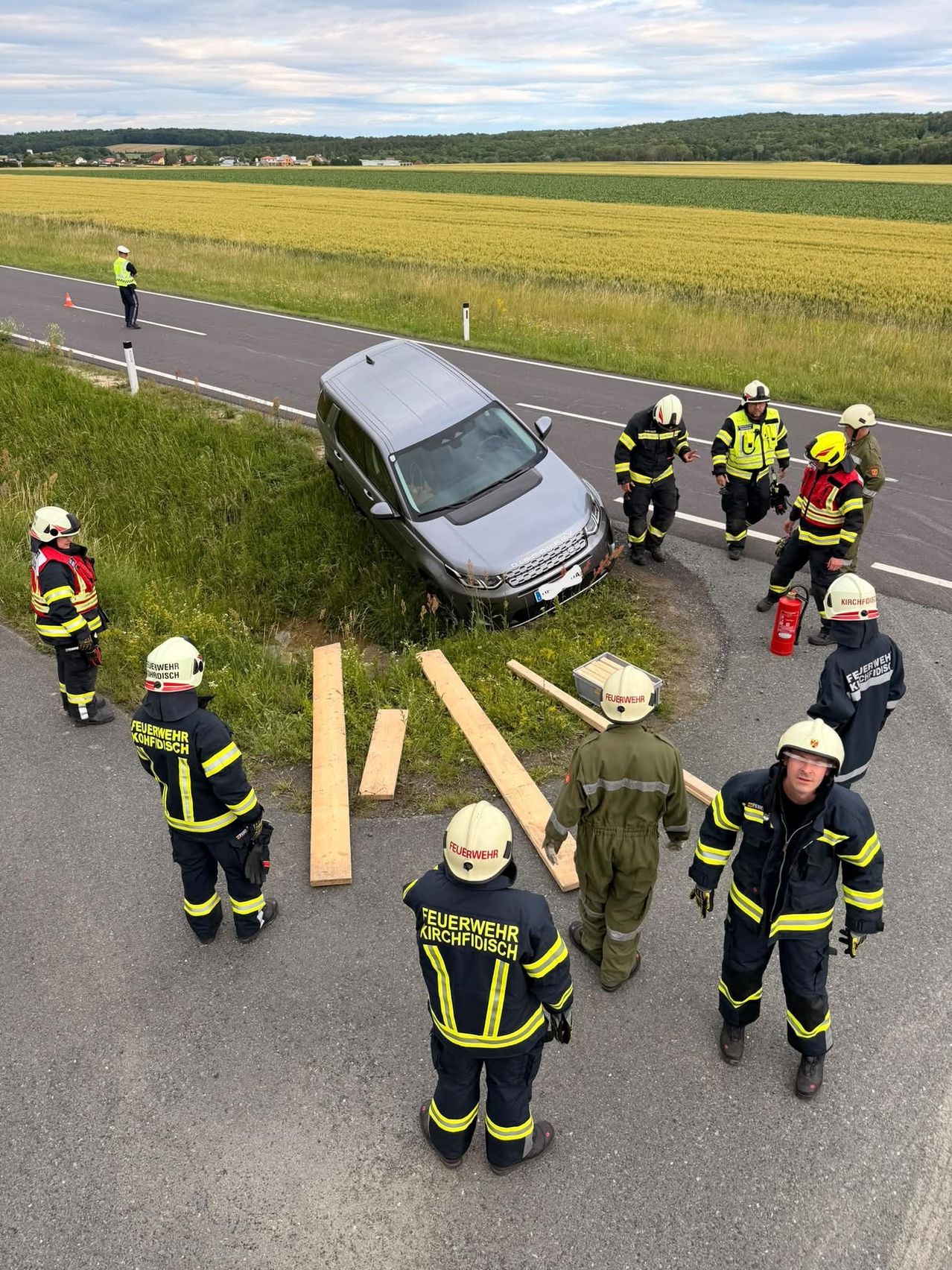 A car is stuck on the side of the road. Firefighters are assessing the situation. Wooden boards are placed around the car for safety.