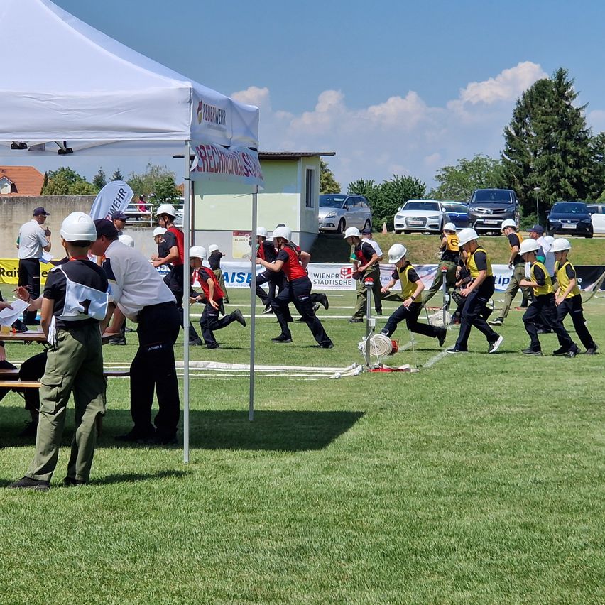 Gruppe von Feuerwehrleuten in Helmen trainiert auf einem Feld mit Zelten und geparkten Autos im Hintergrund.