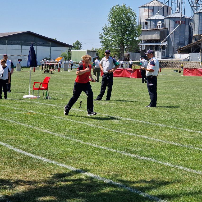Ein Athlet in Rot wirft ein Objekt auf einem Rasenfeld mit weißen Markierungen, während zwei Offizielle zuschauen. Ein roter Stuhl und ein blauer Schirm sind in der Nähe. Gebäude und Bäume sind im Hintergrund.