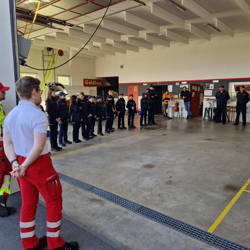 Eine Gruppe von Menschen in Uniformen steht in einer Reihe in einer Feuerwache. Ein Mann in einem weißen Hemd und roten Hosen steht vor ihnen.