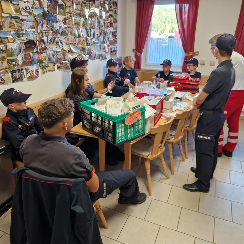 Eine Gruppe von Kindern in Uniform sitzt um einen Tisch herum, möglicherweise in einem Klassenzimmer oder Besprechungsraum. Sie schauen sich die Gegenstände auf dem Tisch an, und ein Erwachsener steht und spricht.