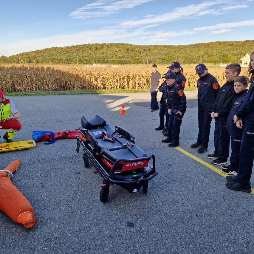 Eine Gruppe von Kindern in Uniformen beobachtet einen medizinischen Wagen und eine Puppe auf dem Boden, mit einer Person in einer reflektierenden Weste, die in der Nähe sitzt, und einem Maisfeld im Hintergrund.