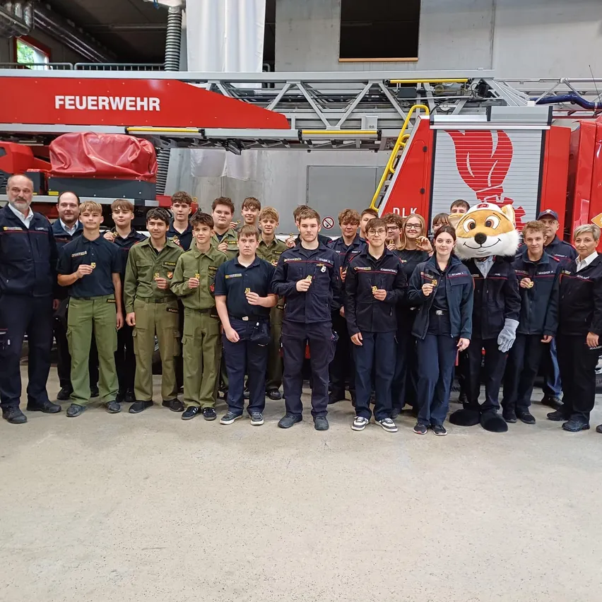 Eine Gruppe von Feuerwehrleuten und jungen Kadetten steht vor einem Feuerwehrauto. Die Feuerwehrleute tragen volle Uniformen, und die Kadetten tragen grüne Uniformen.