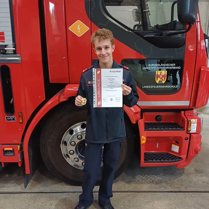 Ein junger Mann in Uniform steht vor einem roten Feuerwehrauto und hält ein Zertifikat.