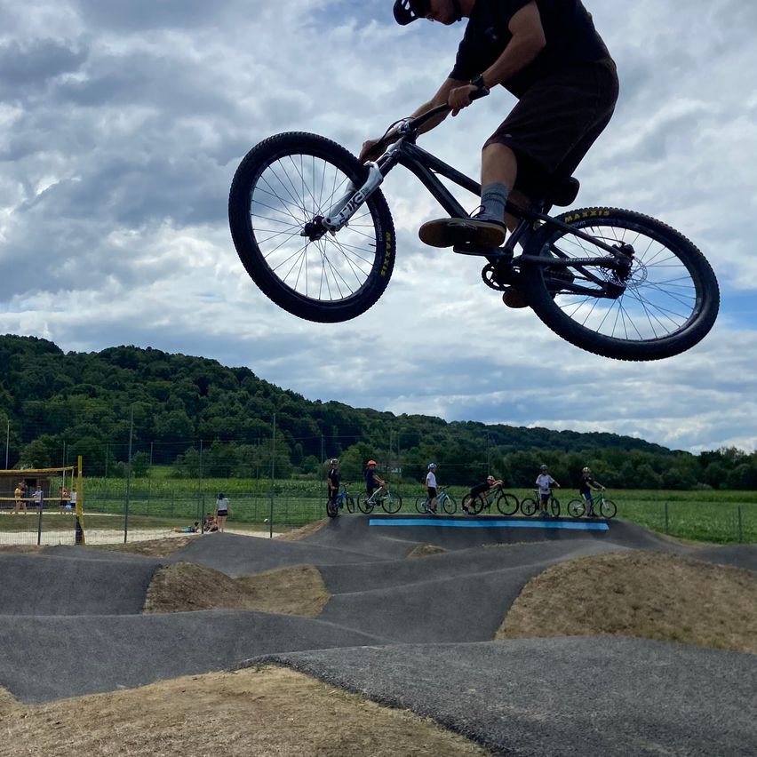 Eine Person führt einen Fahrradtrick in einem Skatepark aus, während andere Radfahrer und ein grünes Feld im Hintergrund zu sehen sind.