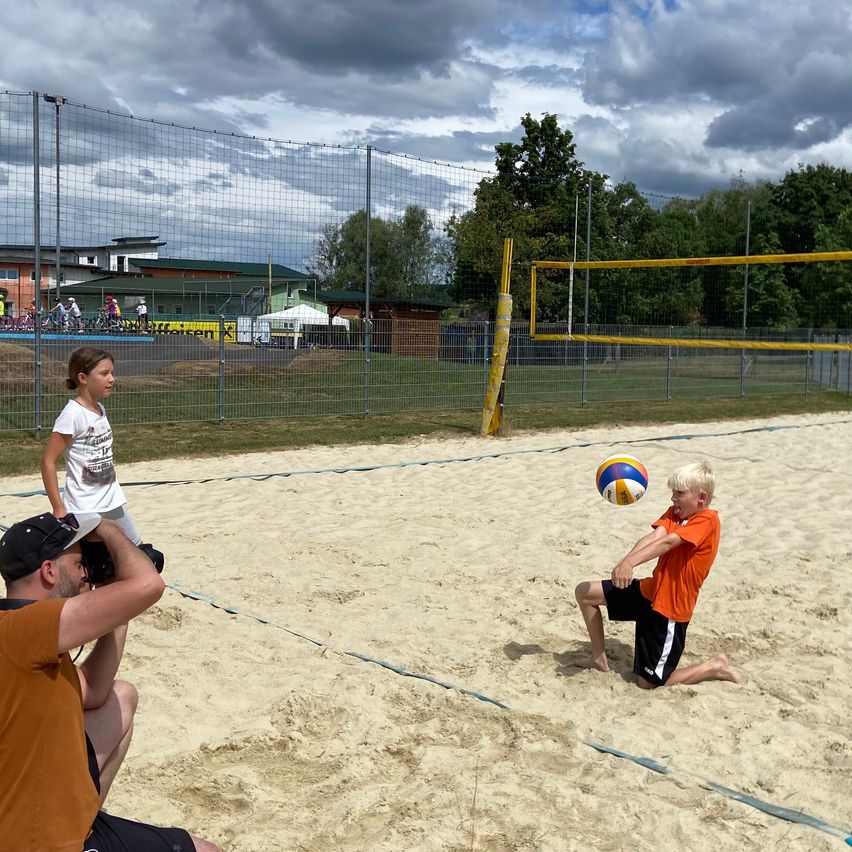 Ein Junge spielt Volleyball auf einem Sandplatz, während ein Mann ein Foto macht. In der Nähe beobachtet ein Mädchen das Spiel. Das Gelände ist von einem Zaun umgeben, mit Bäumen und Gebäuden im Hintergrund.