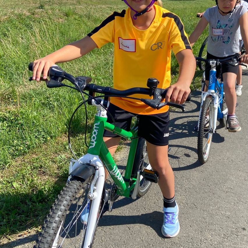 Ein junger Junge fährt ein grünes Fahrrad auf einem Pfad, mit einem anderen Kind, das ein blaues Fahrrad hinter ihm fährt. Sie tragen beide Helme.