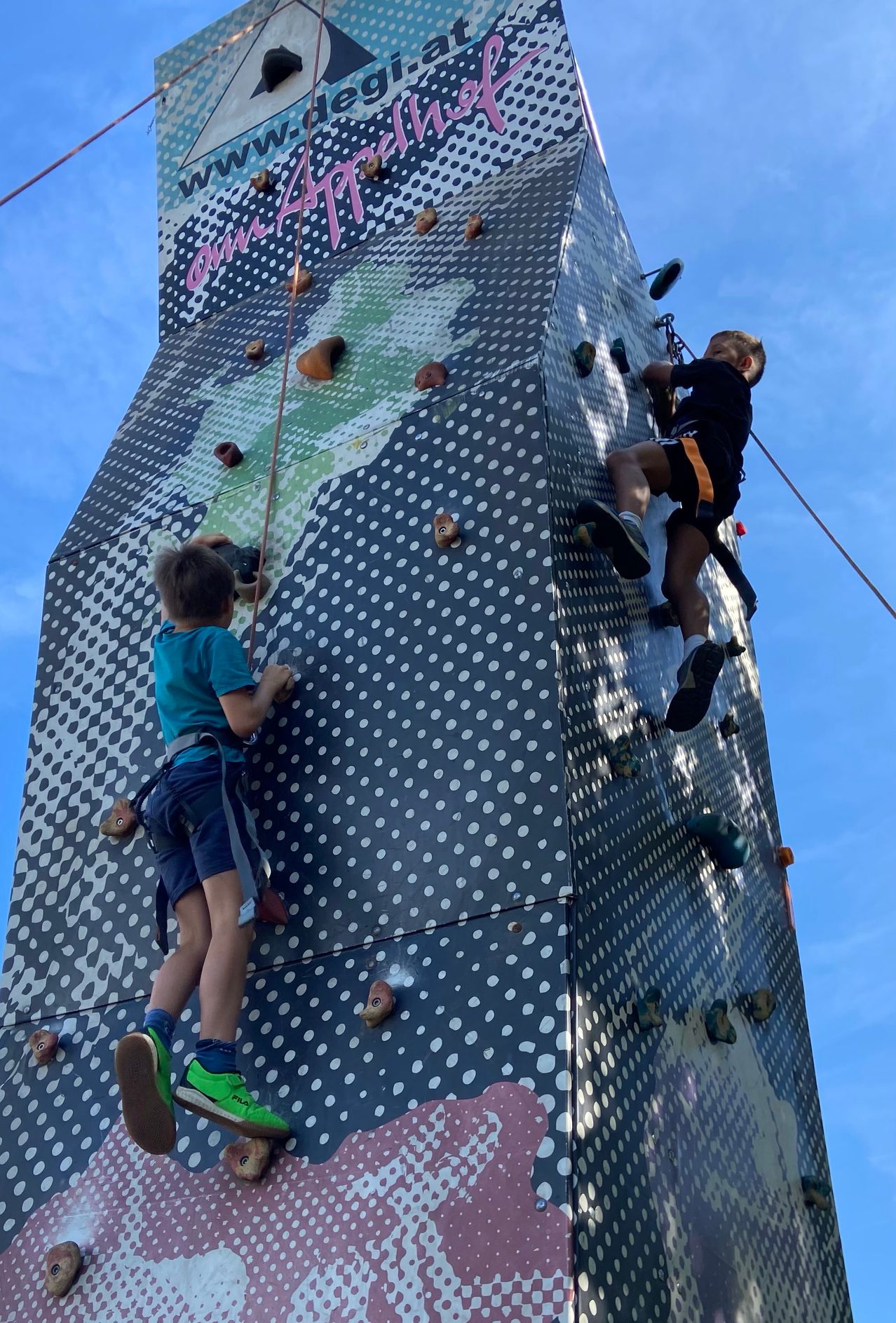 Zwei Kinder klettern an einer Kletterwand. Eines trägt ein grünes Shirt, das andere ein schwarzes. Beide haben Sicherheitsgurte. Die Wand ist mit farbigen Punkten bedeckt.