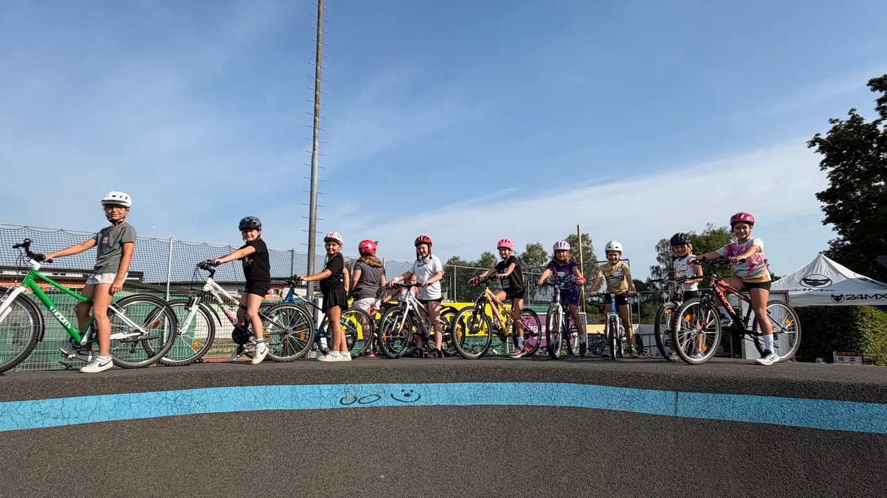 Eine Gruppe junger Radfahrer, alle mit Helmen, steht mit Fahrrädern auf einer Rennstrecke. Die Strecke hat eine blaue Linie an der Seite.