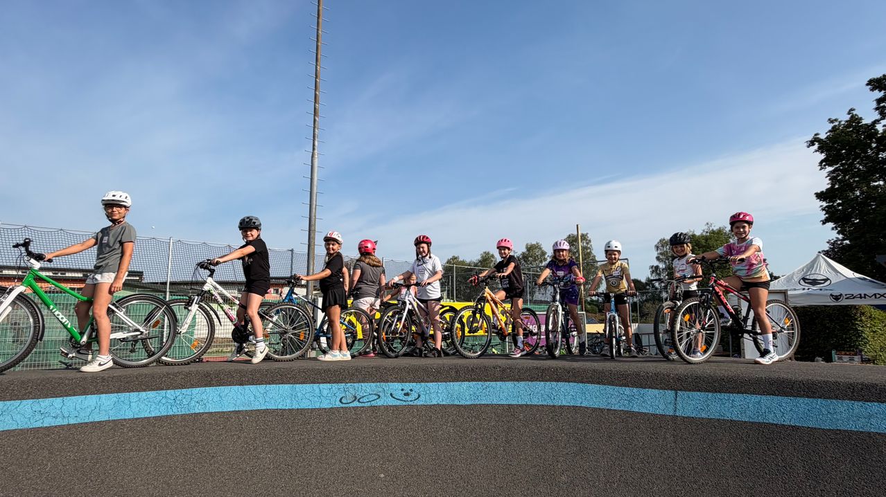 A group of young cyclists, all wearing helmets, are lined up on a track with bikes. The track has a blue line on the side.