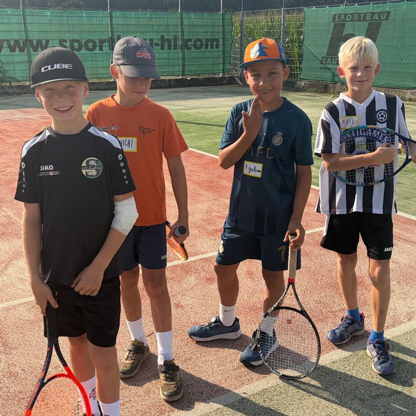 Four boys are standing on a tennis court, each holding a tennis racket. They are wearing different colored jerseys and hats. The boy on the right is wearing a black and white striped shirt. Behind them is a green fence with a banner.
