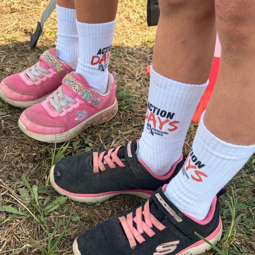 Three children wearing sneakers with colorful socks, one with 'ACTION DAYS' and another with 'MOVE' written on it. They are standing on a grassy field.