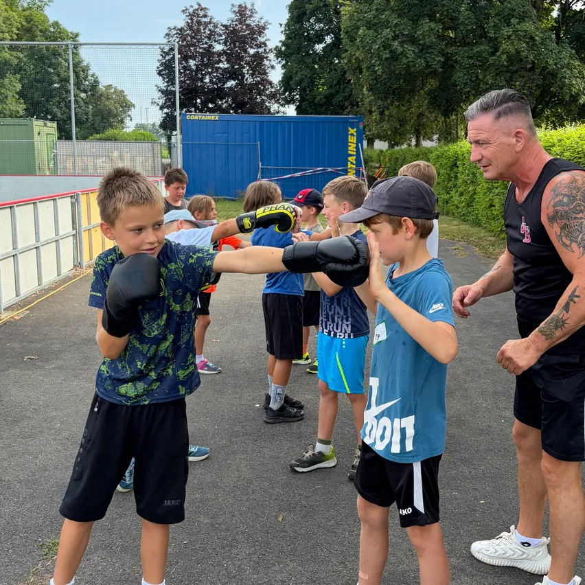 Eine Gruppe junger Jungen in einem Boxkurs, ein Junge mit Handschuhen demonstriert einem anderen Jungen einen Schlag, während ein erwachsener männlicher Trainer aufmerksam zusieht.