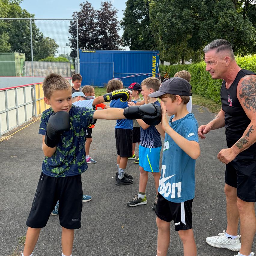 A group of young boys in a boxing class, one boy wearing gloves is demonstrating a punch to another boy, while an adult male coach watches closely.