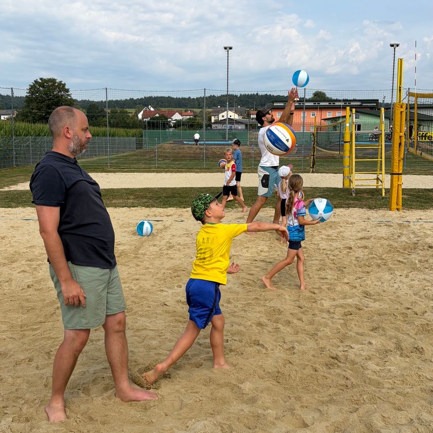 A group of people are playing beach volleyball on a sandy court. A man is jumping to hit the ball while a boy tries to block it.
