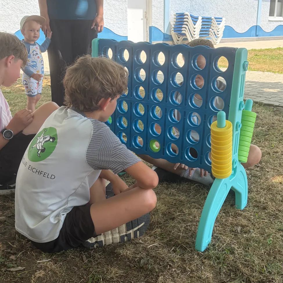 Eine Gruppe von Kindern spielt an einem sonnigen Tag eine Partie Connect Four. Ein Kind trägt ein T-Shirt mit Logo.