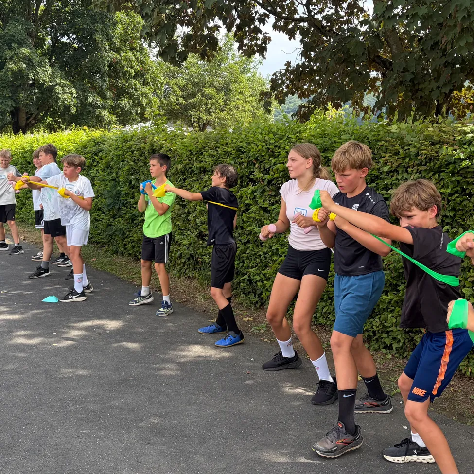 Eine Gruppe von Kindern in Sportkleidung steht auf einem Pfad, hält grüne Widerstandsbänder und scheint an einer Fitnessaktivität teilzunehmen.