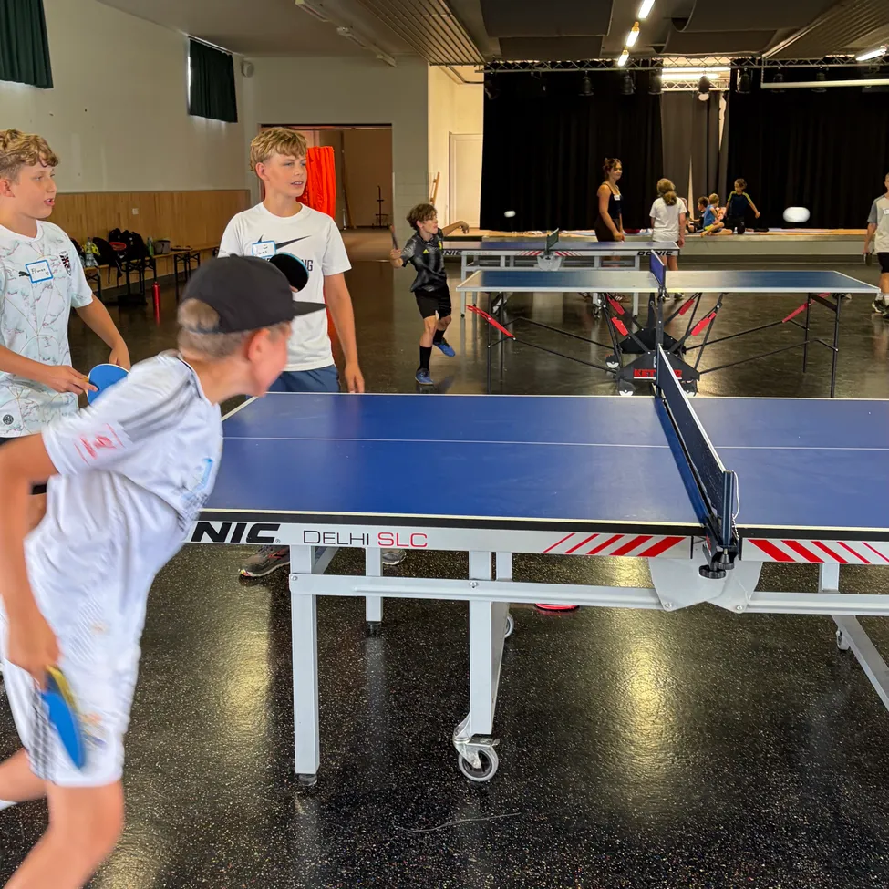 Eine Gruppe junger Jungen spielt Tischtennis in einer Halle. Der Junge im Vordergrund ist dabei, den Ball zu schlagen.