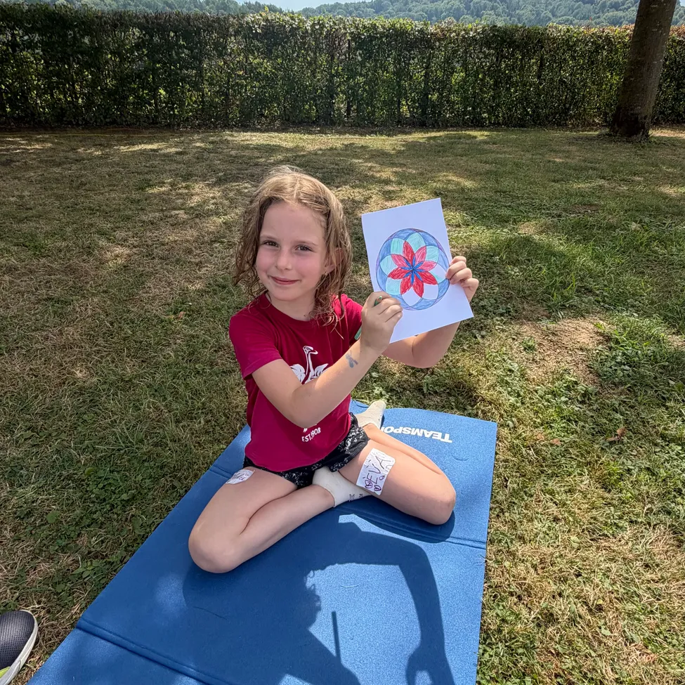 Ein junges Mädchen sitzt auf einer Yogamatte in einem Park und hält eine farbenfrohe blumenförmige Zeichnung hoch.