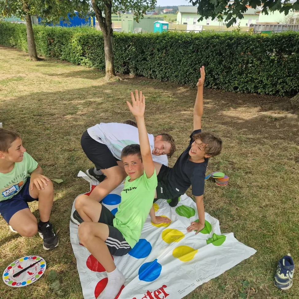 Vier Jungen spielen ein Spiel von Twister auf einem Rasenfeld. Ein Junge liegt auf der Matte, während die anderen darauf stehen.