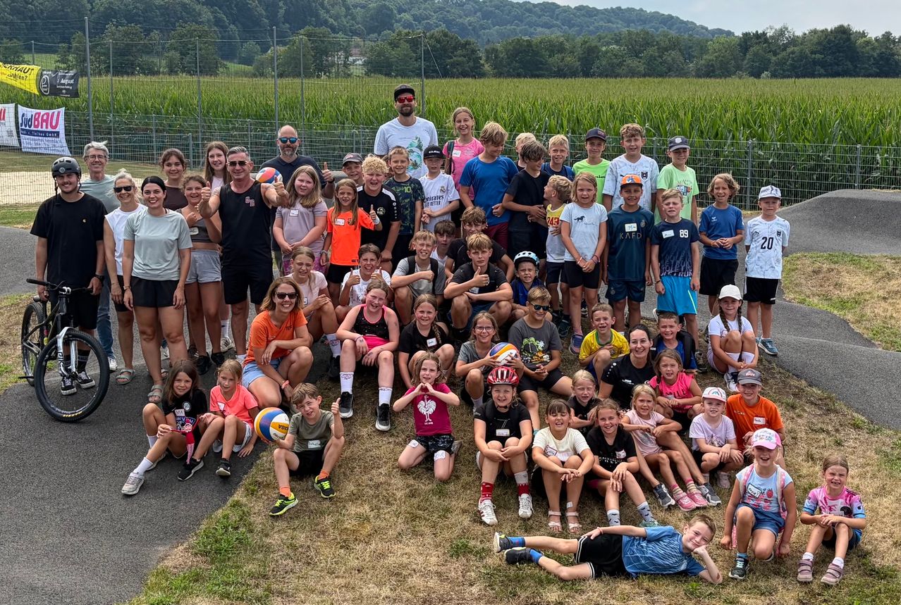 A group of children and adults pose for a photo in a grassy field, some holding sports equipment. They are all smiling and appear to be on a team.