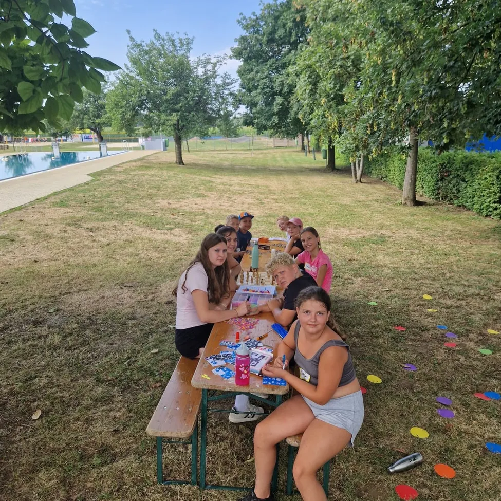 Eine Gruppe von Kindern und Jugendlichen sitzt um einen Picknicktisch in einem Park und spielt Schach. Der Tisch ist mit verschiedenen Gegenständen bestückt, und der Boden ist mit bunten Punkten verziert. Bäume und ein Schwimmbecken sind im Hintergrund zu sehen.