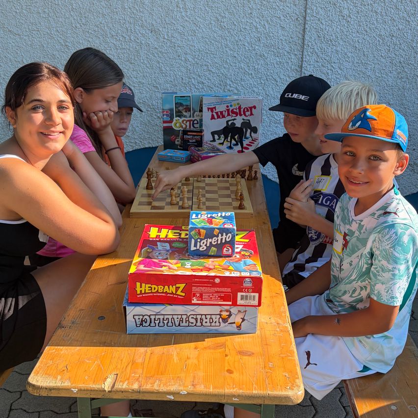A group of children and an adult woman are sitting around a wooden table playing chess and other board games. The woman is smiling and watching the children play.