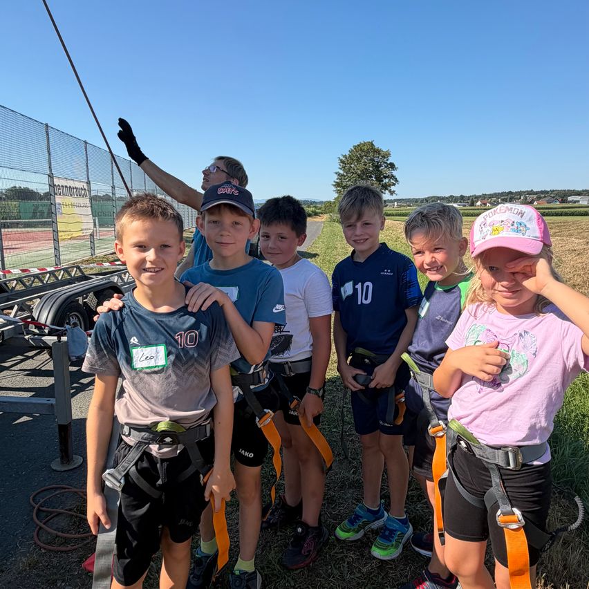 A group of children in outdoor adventure gear pose for a photo on a grassy field with a fence and a tree in the background.