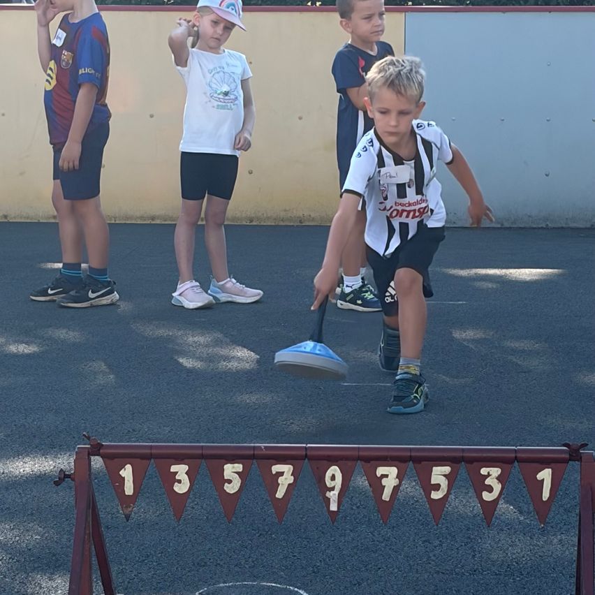 Four boys stand on a playground with numbers on a hurdle in front of them. The boy in the front is about to throw a ball.
