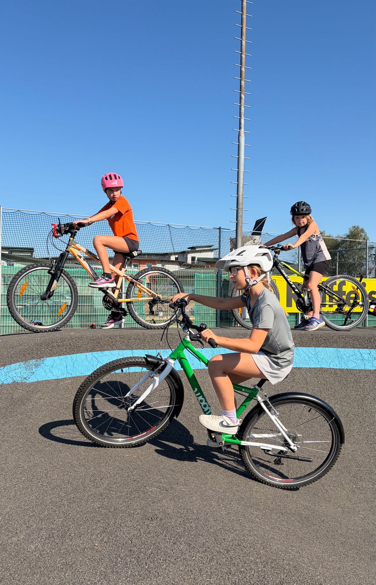 Three young girls ride bicycles on a paved track, wearing helmets and cycling gear. The track is surrounded by a fence and a yellow banner.