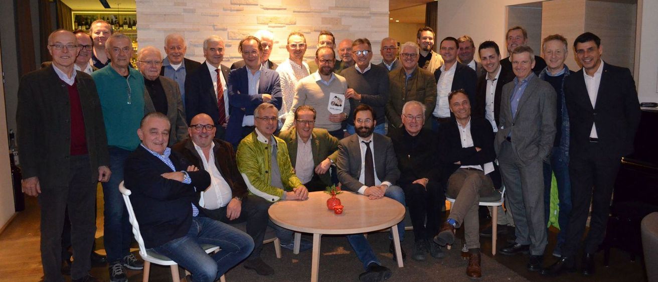 Eine Gruppe älterer Männer, möglicherweise Kollegen, posiert für ein Foto in einem Raum. Einige von ihnen stehen, während andere auf Stühlen sitzen. Sie tragen alle formelle Kleidung. Vor ihnen steht ein Tisch mit einer Blumenvase. Dahinter befindet sich eine Wand mit einer Ziegelsteinstruktur.