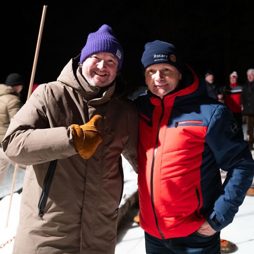 Zwei Männer in Winterkleidung, einer in einem braunen Mantel und einer violetten Mütze, der andere in einer rot-blauen Jacke, lächeln für ein Foto in einem verschneiten Bereich.