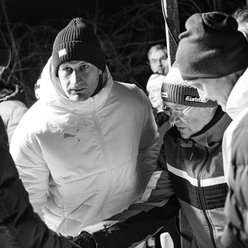 Eine Gruppe von Menschen in Winterkleidung versammelt sich in einem dunklen Bereich. Der Mann in der Mitte schaut nach vorne. Die Person rechts trägt eine Mütze mit der Aufschrift 'Eisbo'. Die Person links trägt eine schwarze Jacke.