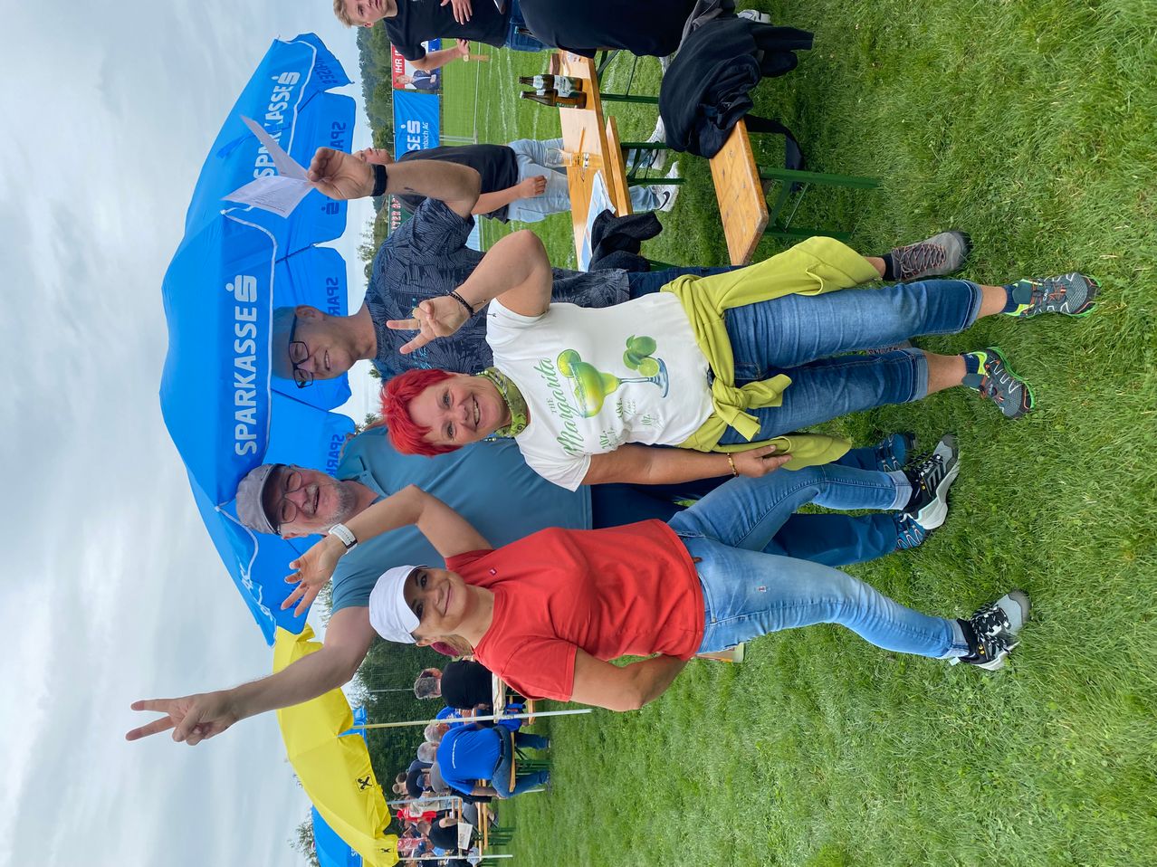 Mehrere Personen posieren für ein Foto auf einem Rasenfeld unter einem blauen Schirm, mit einem verschwommenen Hintergrund von sitzenden und stehenden Personen.