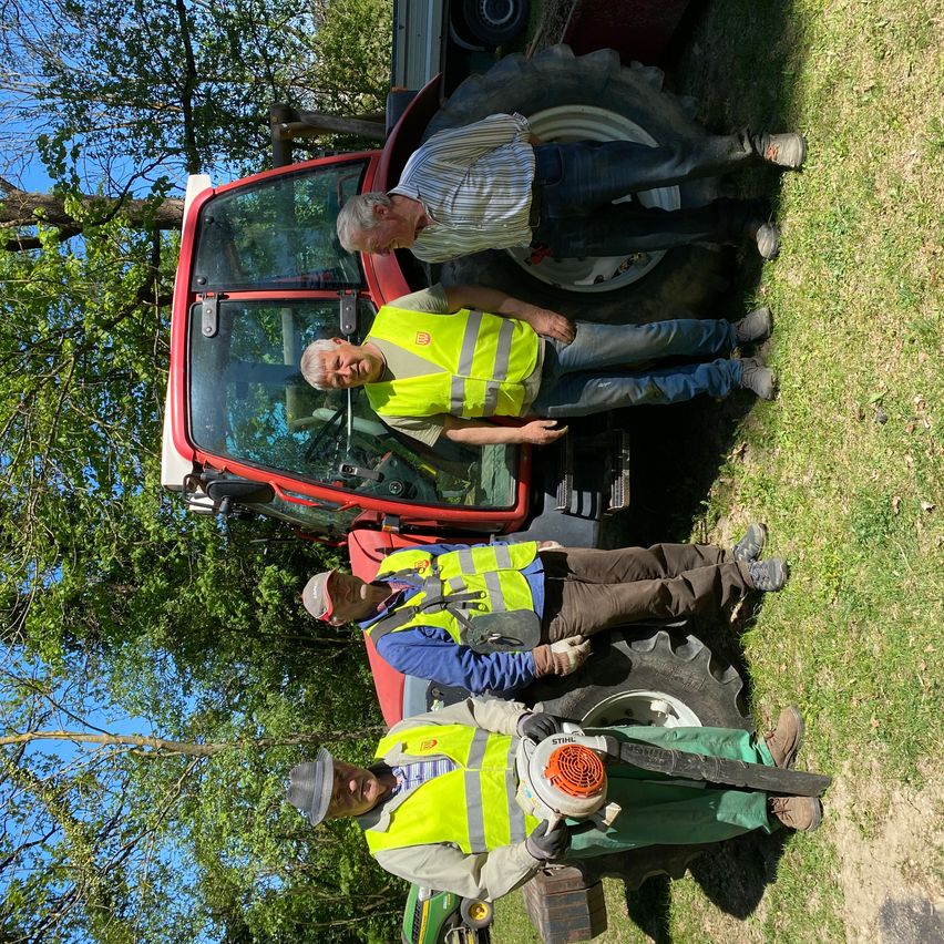 Drei Männer in gelben Warnwesten posieren für ein Foto vor einem roten Traktor auf einem Grasfeld. Bäume sind im Hintergrund zu sehen.